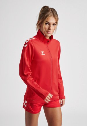 Red athletic jacket with a high collar, zip closure, and side pockets. Features white chevron accents on sleeves and branding on the chest.