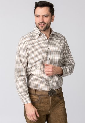 Mann trägt beige-weiß kariertes Hemd und braune Lederhose mit dekorativer Gürtelschnalle, posiert vor einem hellgrauen Hintergrund.