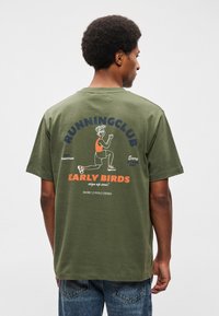 T-shirt de algodão verde-azeitona, apresentando um design gráfico nas costas com texto e uma ilustração de um corredor. Mangas curtas, corte descontraído.