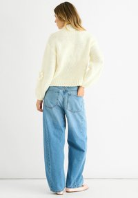Woman standing sideways with hand in back pocket, wearing a cream knit sweater and wide-legged blue jeans against white background.