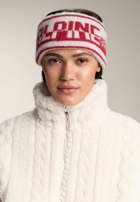 Bentita tricotată roșie și albă cu text îndrăzneț „ALPINE”, având dungi orizontale. Purtați cu o jachetă albă din fleece texturat.