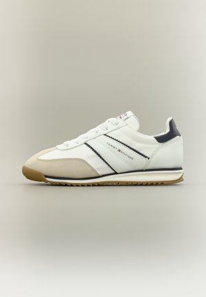 Baskets de sport blanches avec une tige en cuir lisse, une semelle en caoutchouc gomme, des rayures noires et un petit détail de logo sur le côté.