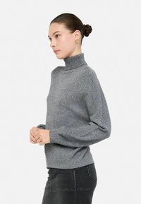 Grauer Strickpullover mit hohem Kragen, lockerem Schnitt, strukturierten Oberflächen und gerippten Bündchen. Das Model trägt schwarze Jeans und zeigt das Profil von der Seite.