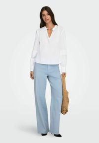 Femme portant un chemisier blanc à manches longues et un pantalon large bleu clair, tenant un sac fourre-tout tissé marron, debout sur un fond uni.