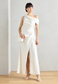 White, one-shoulder gown with draped fabric, side slit, and soft texture. Paired with a small, white crossbody bag and strappy heels.