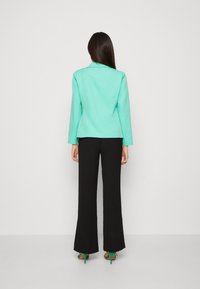 Kvinna med rakt långt hår iförd en mintgrön kavaj, svarta vida byxor och gröna högklackade sandaler, vänd bortåt mot vit bakgrund.