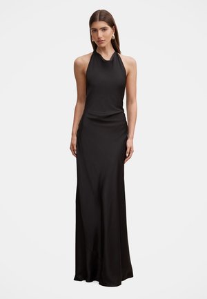 Femme debout portant une longue robe noire sans manches à col halter avec des accessoires minimalistes sur fond blanc uni.