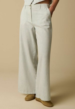 Pantalon large couleur crème avec fines rayures verticales, porté avec des chaussures marron perforées type slip-on, sur un fond beige.