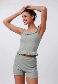 Mladá žena se usmívá, má ruce zdvižené a na sobě má odpovídající světle šedou zalichrovanou soupravu s krátkým topem a šortkami s volanty.