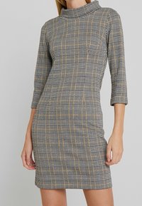 Abito aderente con colletto alto, caratterizzato da un motivo a quadretti in bianco, nero e giallo. Realizzato in materiale elasticizzato con maniche tre quarti.
