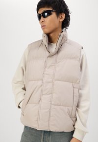 Gevoerde vest in lichtbeige met hoge kraag, voorzakken en een zachte textuur. Draag het over een lichte trui, aangevuld met donkere zonnebrillen.