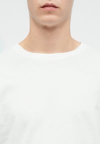 Den nederste del af ansigtet og nakken på en person iført en simpel hvid T-shirt med rund hals mod en hvid baggrund.