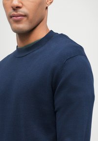 Granatowy sweter w miękkim materiale z okrągłym dekoltem. Posiada ściągacze przy mankietach i dolnym końcu, ma gładką teksturę i brak widocznych wzorów.