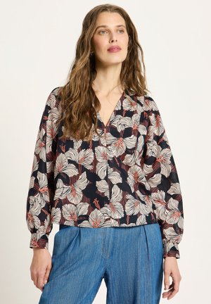 Woman with long wavy hair wearing a black blouse with large white and red floral patterns and loose blue jeans, standing against a plain background.