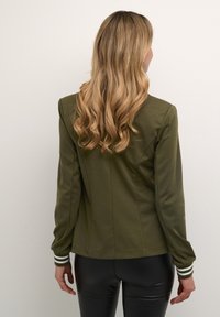 Olivgrön, åtsittande jacka med en slät yta, kontrasterande vitrandiga manschetter och strukturerad design. Bärs med svarta leggings.