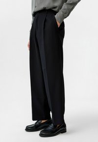 Zwarte geplooide broeken met een brede Silhouette en verticale streepjes, gecombineerd met zwarte instappers, gepresenteerd tegen een neutrale achtergrond.