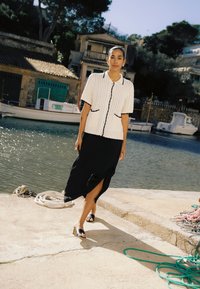 Femme marchant sur un quai au bord de l'eau, portant un cardigan en maille blanc à manches courtes, une jupe midi noire fendue et des sandales noires, près de bateaux et de cordes enroulées.