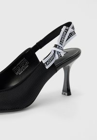 Černý slingback podpatek s vroubkovanou texturou, špičatou špičkou a lesklým podpatkem. Obsahuje bílý textilní pásek s černým logem.