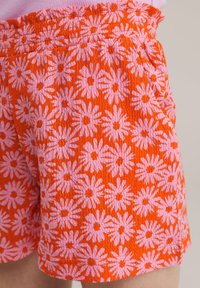 Oranžne kratke hlače s rožnato cvetličnim vzorcem, teksturirana tkanina, elastičen pas in stranski žepi. Oblikovanje vključuje cvetove, podobne margaretam.