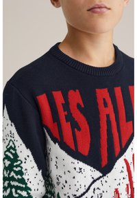 Navy zils adīts džemperis ar sarkanu tekstu "LES ALPES", baltām sniega klātiem kalniem un zaļām kokiem rakstainajā dizainā.