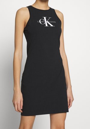 Černé šaty bez rukávů s bílým logem „Calvin Klein Jeans“ na přední straně, nošené osobou s pažemi volně podél těla.