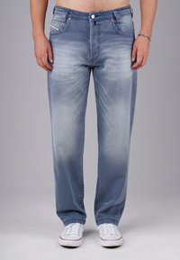 Man wearing faded blue jeans and white sneakers, standing with arms relaxed by sides against a plain gray background.