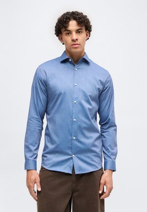 Joven con cabello rizado que lleva una camisa azul claro de manga larga abotonada y pantalones marrones, de pie frente a un fondo blanco liso.