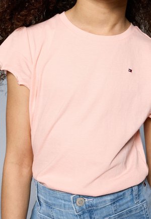 Jong persoon met krullend haar, gekleed in een lichtroze T-shirt met korte mouwen en lichtblauwe high-waisted spijkerbroek.