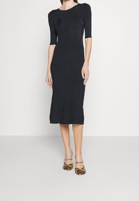 Robe noire ajustée avec des manches courtes, texture côtelée et jupe arrivant au genou. Associée à des talons à motifs avec des accents serpentin.