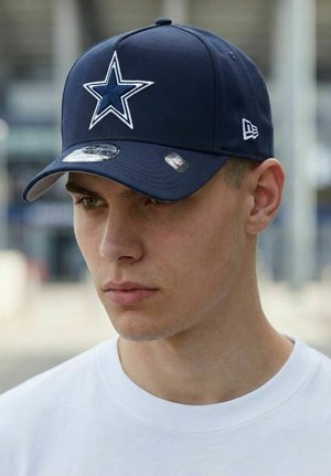 Young man wearing a navy blue baseball cap with a white star logo and a white shirt, outdoors in an urban setting.