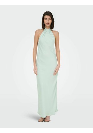 Femme aux cheveux bruns mi-longs, vêtue d'une robe maxi sans manches vert pâle à col licou et de sandales beiges, debout devant un fond uni.