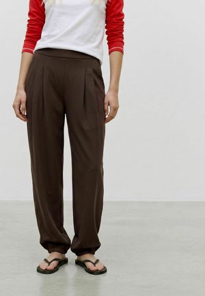 Femme portant un pantalon plissé marron avec des poignets élastiques, associé à une chemise blanche et rouge à manches longues et des sandales noires.