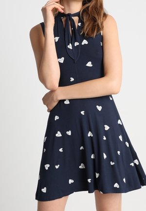 Robe sans manches bleu marine à motifs de cœurs blancs, avec un détail noué au col et une jupe évasée.