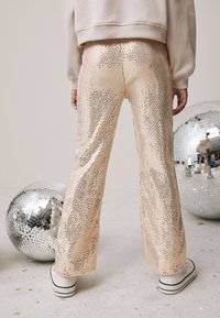 Pantaloni realizzati in paillettes metalliche lucide di tonalità pesca, con silhouette svasata e superficie testurizzata, abbinati a sneakers bianche.