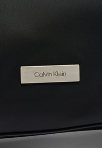 Ορθογώνια ασημί μεταλλική ετικέτα με την επιγραφή "Calvin Klein" προσαρτημένη σε μαύρη υφή υφάσματος.