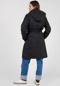 Zwarte gewatteerde jas met een capuchon, een getailleerde taille en een middellange lengte. Gecombineerd met blauwe spijkerbroek met opgerolde manchetten en witte sneakers.