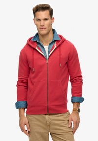 Felpa rossa con zip realizzata in morbido tessuto, dotata di una tasca frontale, cordini regolabili e una camicia di denim indossata sotto.