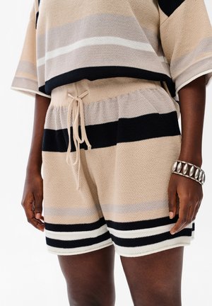 Dzianinowe beżowe, czarne i białe szorty w paski z sznurkiem w pasie, noszone z dopasowanym krótkim topem i srebrną bransoletką na prawym nadgarstku.