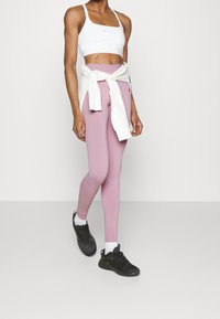 Kvinna klädd i vit sportbh, ljusrosa leggings, vit sweatshirt knuten i midjan, vita strumpor och svarta löparskor som går på en enfärgad bakgrund.