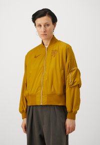 Jachetă galbenă tip bomber cu fermoar în față, mansete ribbed și un grafic cu leu. Are un buzunar lateral cu fermoar și un material fin, ușor.