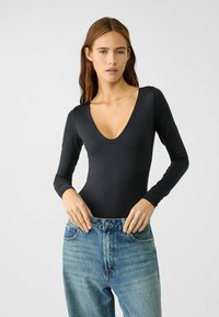 Schwarzer Langarm-Bodysuit mit tiefem V-Ausschnitt. Aus glattem Material, figurbetontes Design, kombiniert mit hochgeschnittenen blauen Jeans.