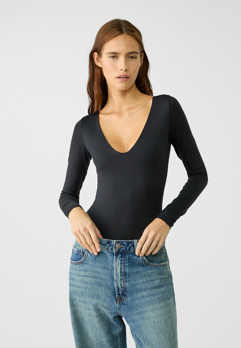 Schwarzer Langarm-Bodysuit mit tiefem V-Ausschnitt. Aus glattem Material, figurbetontes Design, kombiniert mit hochgeschnittenen blauen Jeans.