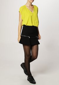Blouse jaune à manches courtes avec un décolleté en V, associée à une mini-jupe noire avec des poches zippées, portée avec des chaussures noires à clous et des bas.