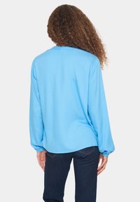 Camisa de manga larga azul claro con un patrón texturizado y puños elásticos, combinada con vaqueros oscuros. La parte trasera presenta un diseño de corte holgado.