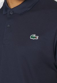 Närbild av en marinblå polo med två knappar och en grön krokodil-logotyp broderad på bröstet.