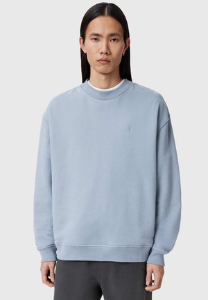 Junge Person mit langen dunklen Haaren trägt ein hellblaues Rundhals-Sweatshirt und dunkle Hose, steht vor hellem grauem Hintergrund.