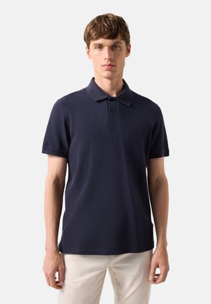 Jonge man die een marineblauw poloshirt met korte mouwen en lichtbeige broek draagt, staand tegen een effen lichtgrijze achtergrond.