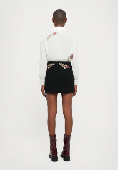 White blouse with floral embroidery on the shoulder; black mini skirt with embroidered floral accents. Model wearing glossy, burgundy ankle boots.