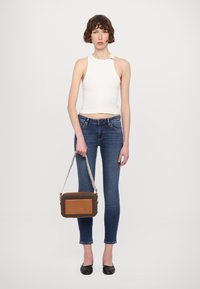 Borsa a tracolla marrone e a fantasia con pannello frontale liscio, hardware in argento e tracolla staccabile, indossata da un modello con jeans aderenti.