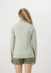 Hellgrüne Fleecejacke mit hohem Kragen und langen Ärmeln, die eine glatte Textur und eine taillierte Passform aufweist. Darunter werden kurze olivgrüne Shorts getragen.